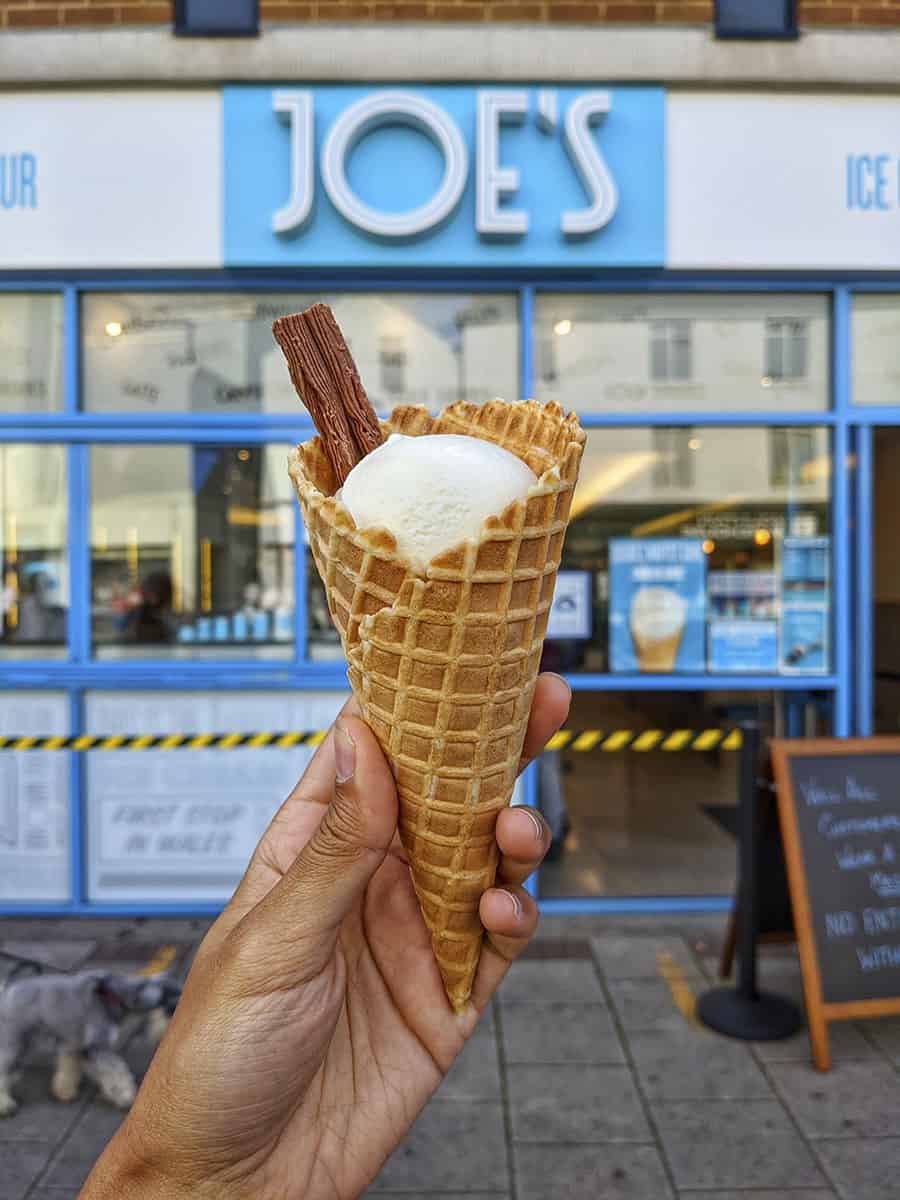 An ice cream cone with vanilla ice cream and a flake held up in front of a shop called Joe's