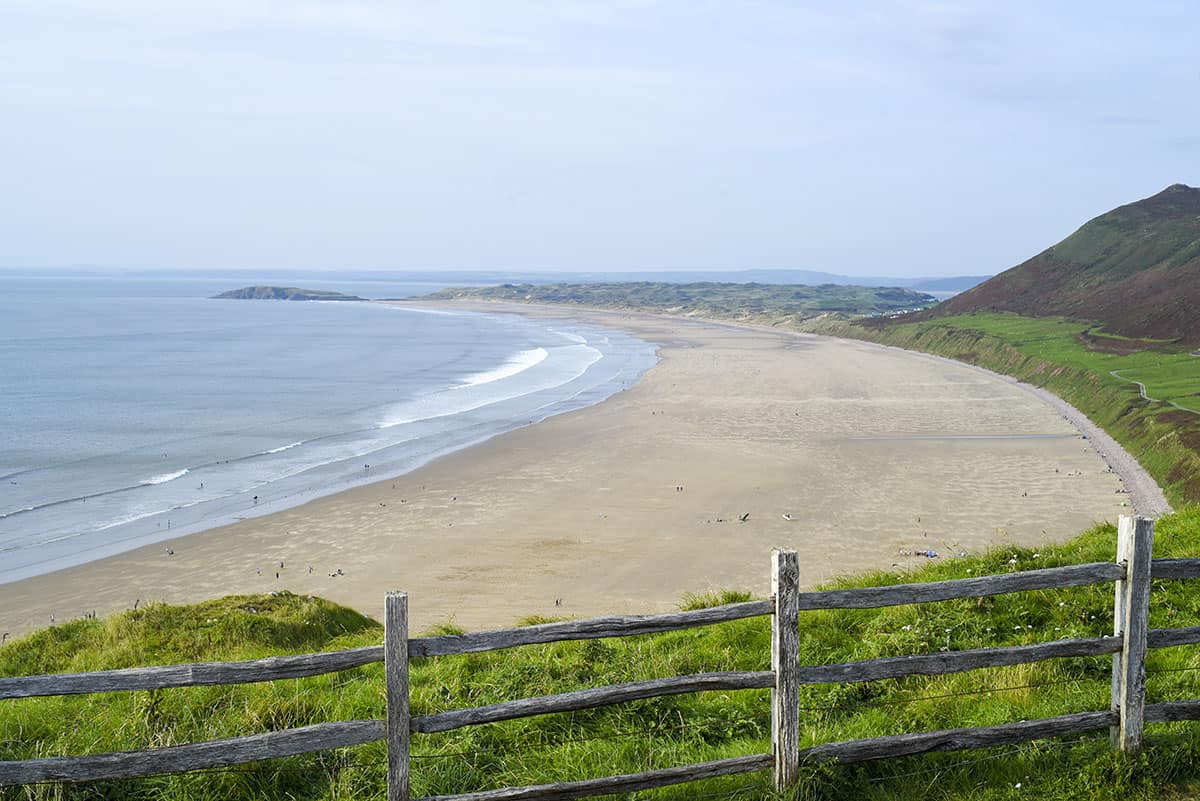 A wide view of a vast sandy beach with grass and a wooden fence in the foreground