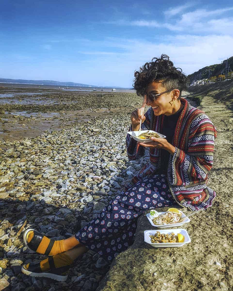 A girl sitting on a pebble beach eating takeaway seafood