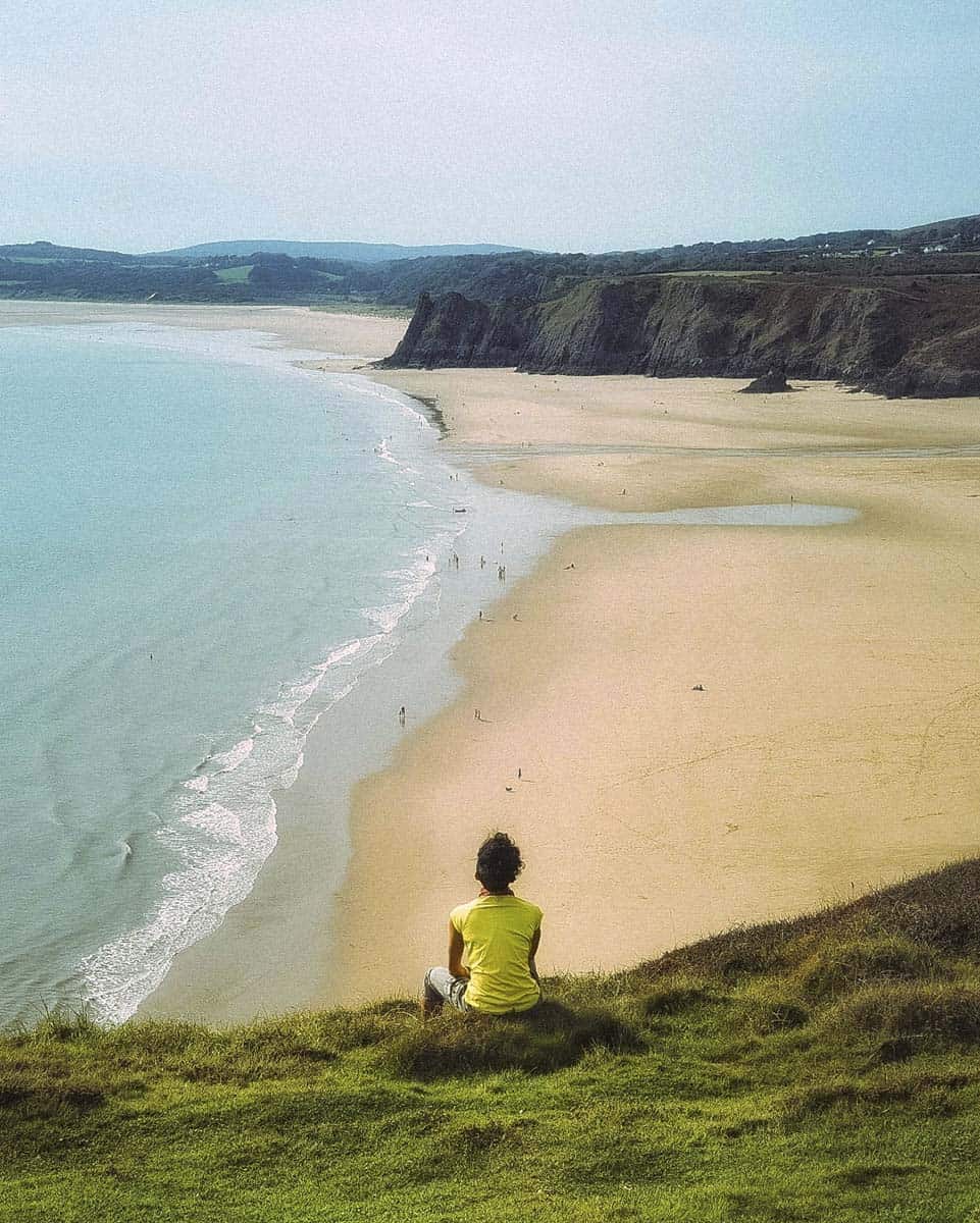 A girl sitting on a grass mound on a clifftop looking out over a beautiful beach