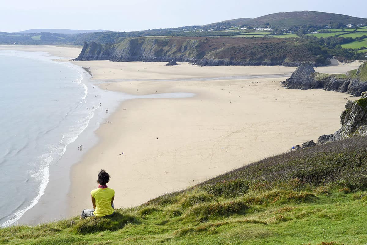 A woman in a yellow top on a grassy cliff top bank looking out over Three Cliffs Bay in Gower, Wales