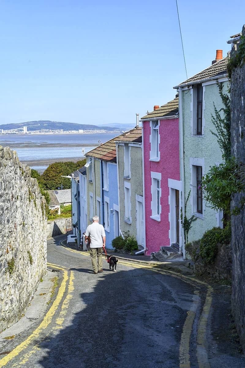 A man walking a dog down a steep lane lined with colourful pastel houses