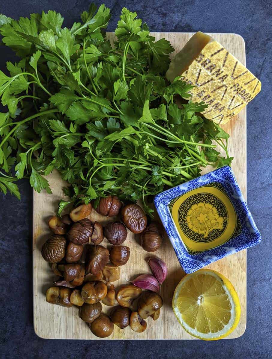 A wooden chopping board viewed from above, laden with vibrant green parsley, cooked chestnuts, a big hunk of Grana Padano cheese, a small ceramic dish containing oil, and half a lemon