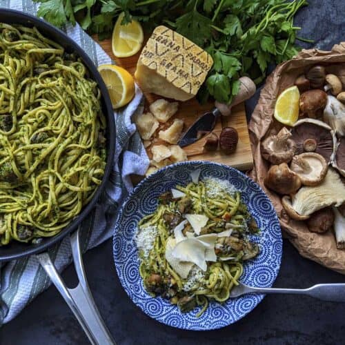 A frying pan full of a pasta dish that's just been cooked, set down on a tea towel on a wooden board, alongside a pretty blue bowl containing the pasta plated up, as well as the raw ingredients that went into the pasta dish, including parsley, Grana Padano cheese, mushrooms, chestnuts and lemon