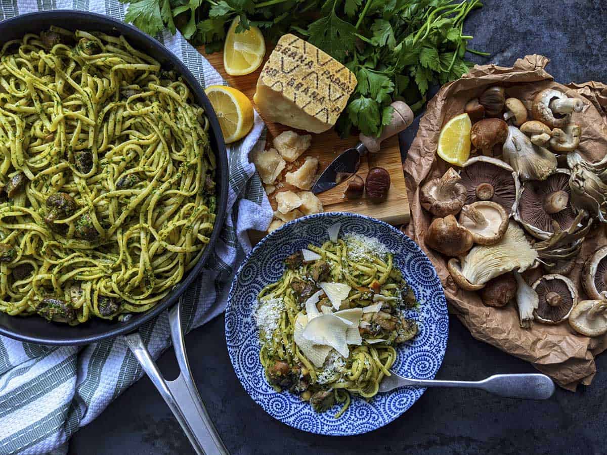 A frying pan full of a just-cooked pasta dish, alongside a bowl of plated up pasta and the raw ingredients within the dish, including Grana Padano cheese, mushrooms, lemon, parsley and chestnuts.