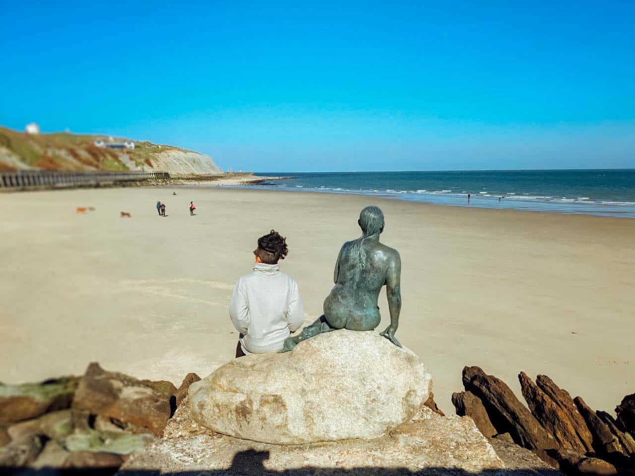 A girl sitting on a rock next to a sulpture of a naked woman, both looking out over a sandy beach towards the sea under a blue sky