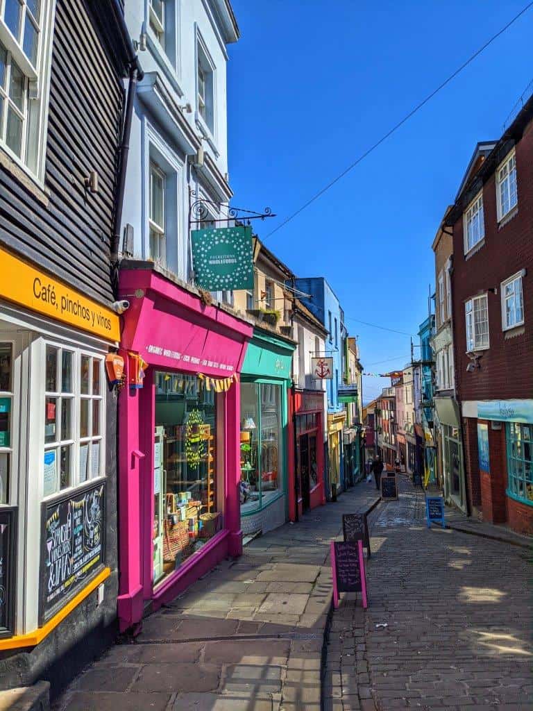 A view down a narrow cobbled street lined with colourful independent shop and restaurant fronts with chalk boards out the fronts under a blue sky