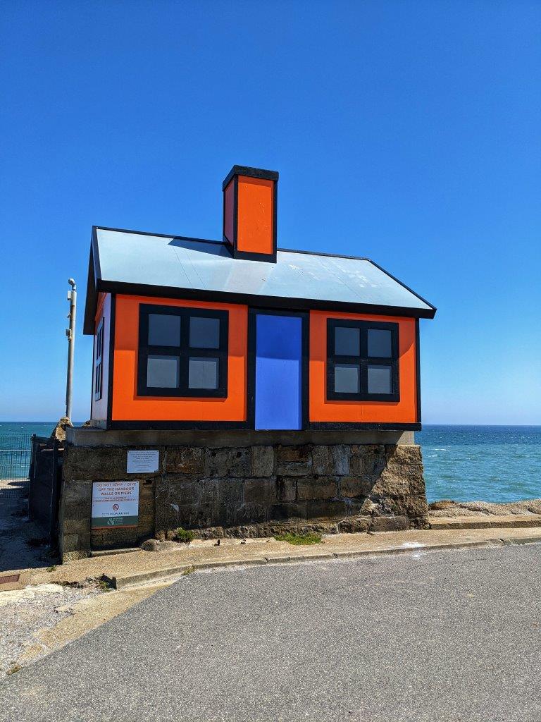 A mock house, one third of the size of a real house, of bright orange with a bright blue door against the background of a blue sea