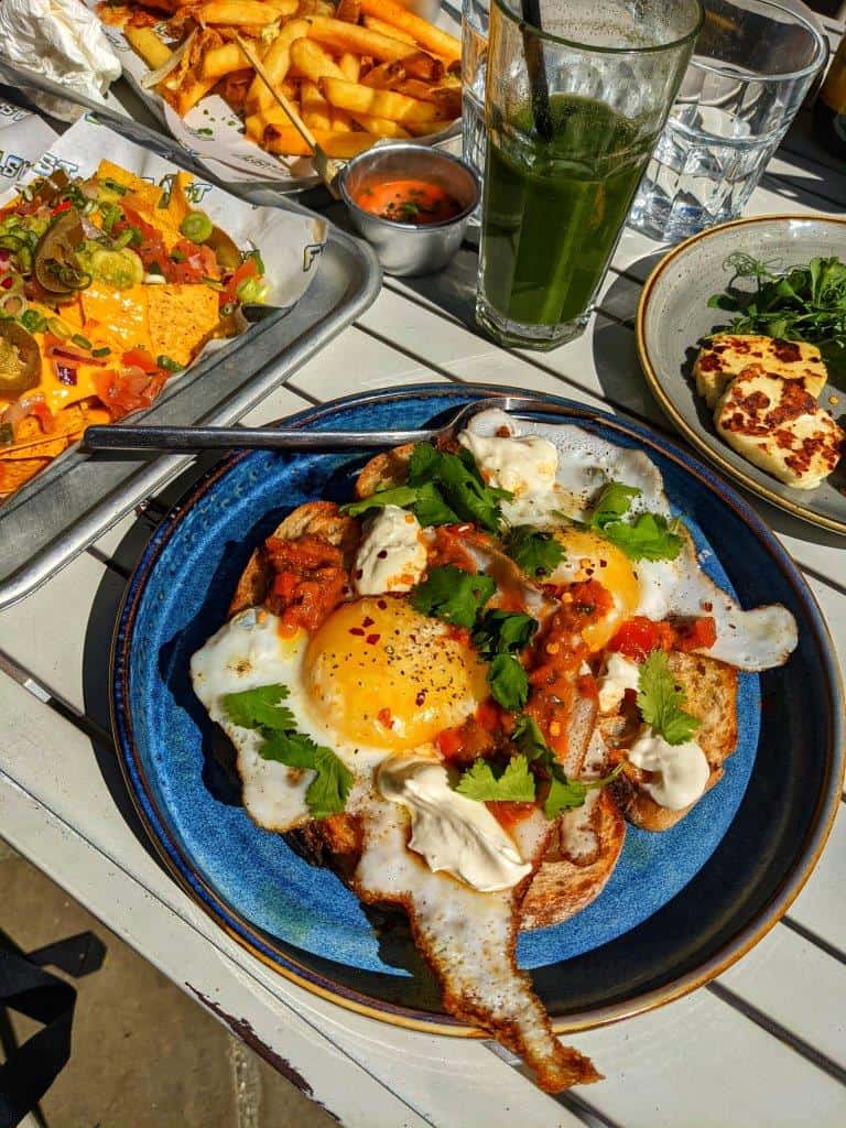 A restaurant dish of two large duck eggs on sourdough toast topped with coriander and tomato chutney, with a side of grilled halloumi, next to a loaded plate of nachos