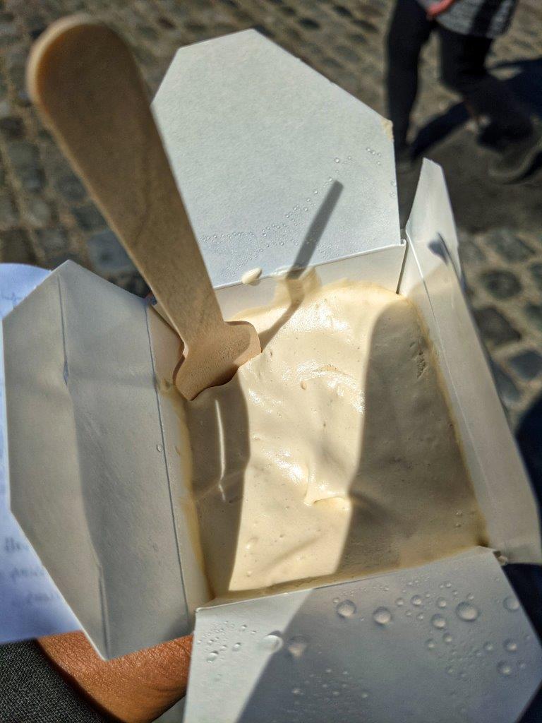 A small square cardboard container with the lid open to reveal ice cream inside with a wooden spoon sticking out the top