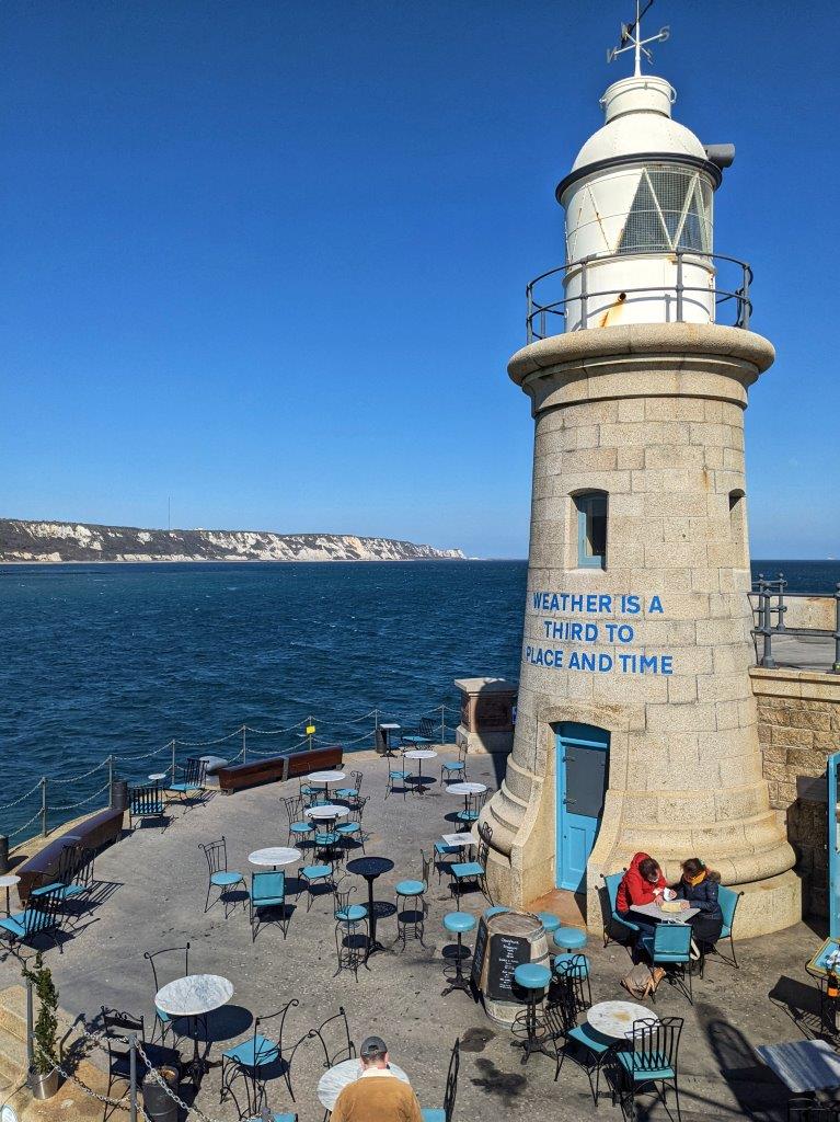 A small lighthouse on the end of the harbour arm with seats around its entrance and blue sea and skies in the background