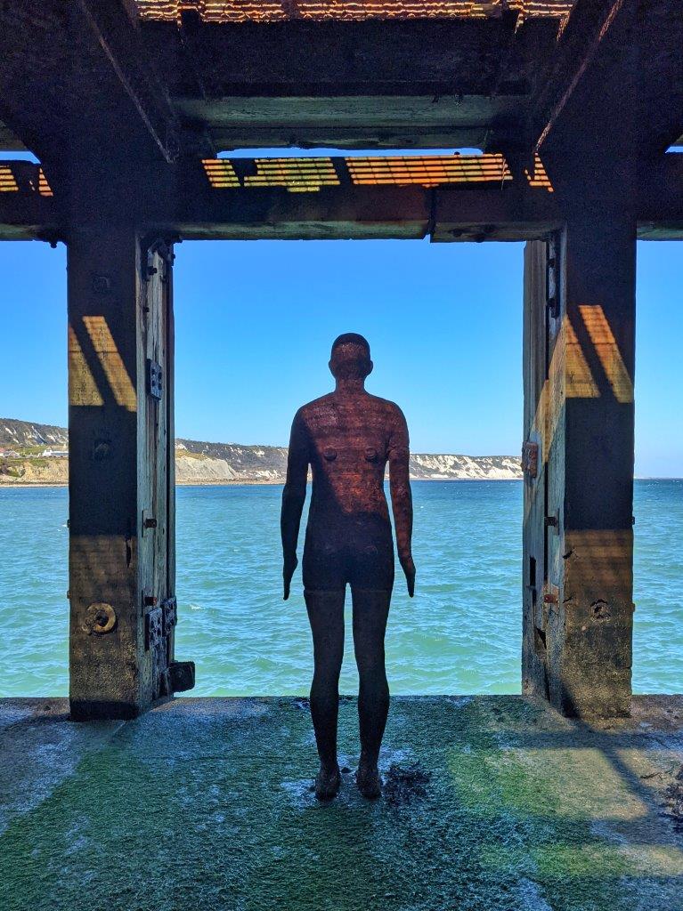 A lone and haunting scuputre of an iron man under a harbour arm looking out over a blue sea