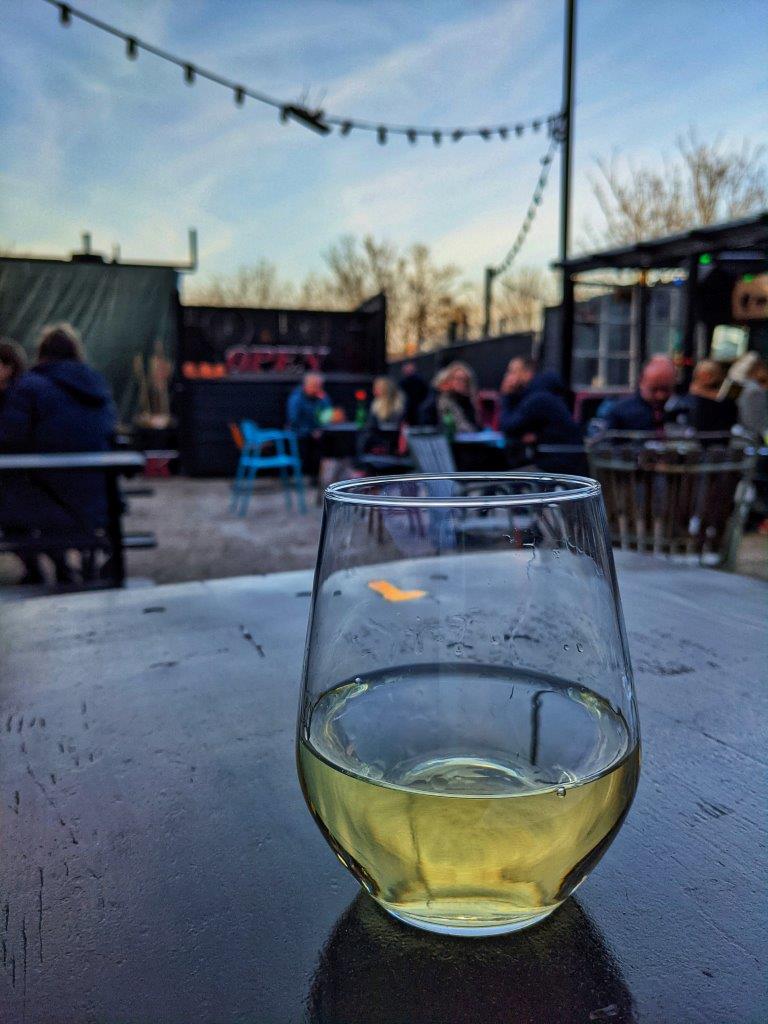 A glass of wine set on a table outside against a backdrop of patrons sitting at their own tables under at dusk