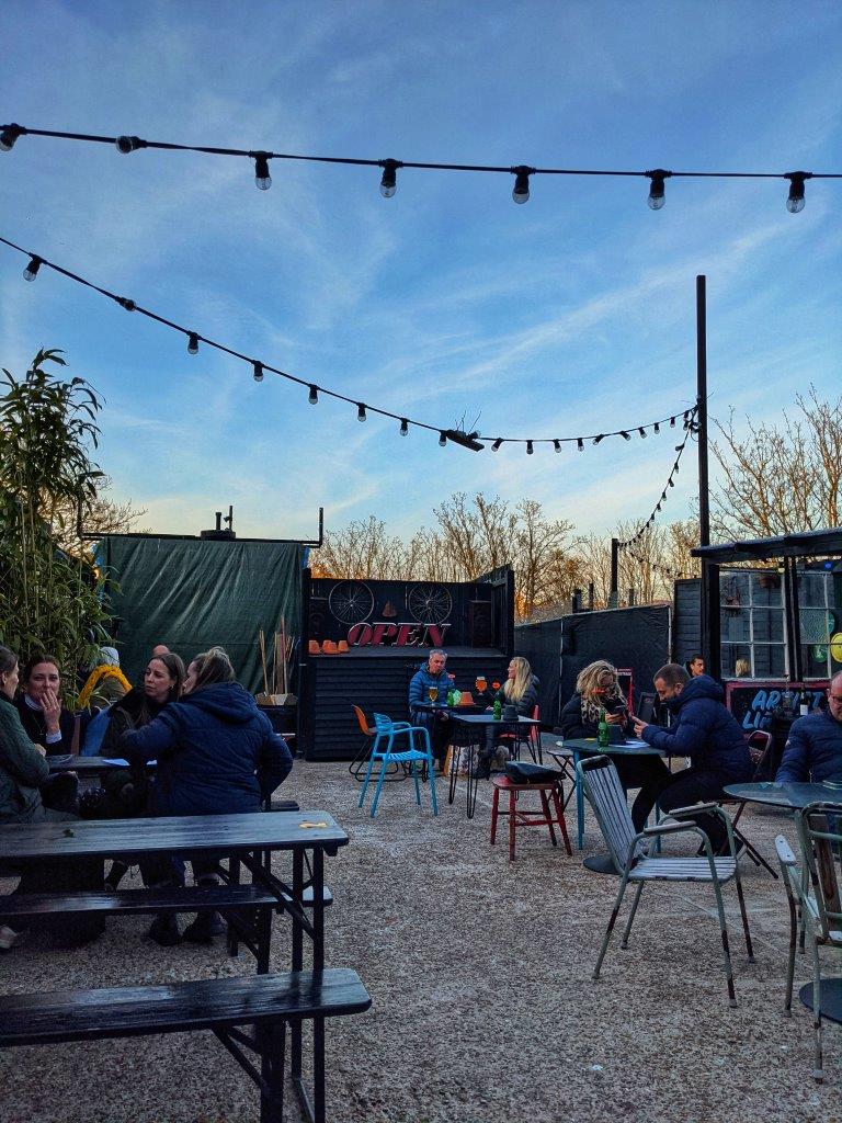 Picnic tables and small metal tables occupied by customers wrapped up in winter coats drinking beverages under a dusk sky