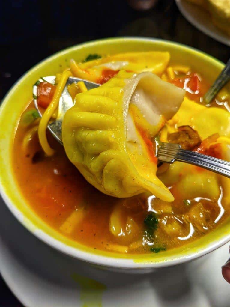 A close up of a pleated crescent-shaped dumpling on a spoon being lifted out of a turmeric-stained noodle soup