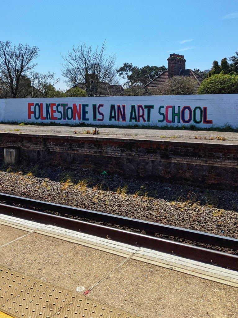 The words 'Folkestone Is An Art School' in large letters painted onto the side of a wall running the length of some railway tracks