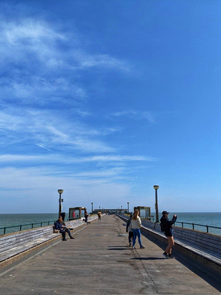 Lots of blue sky and a view looking down a pier with a few people walking along, the sea in the distance