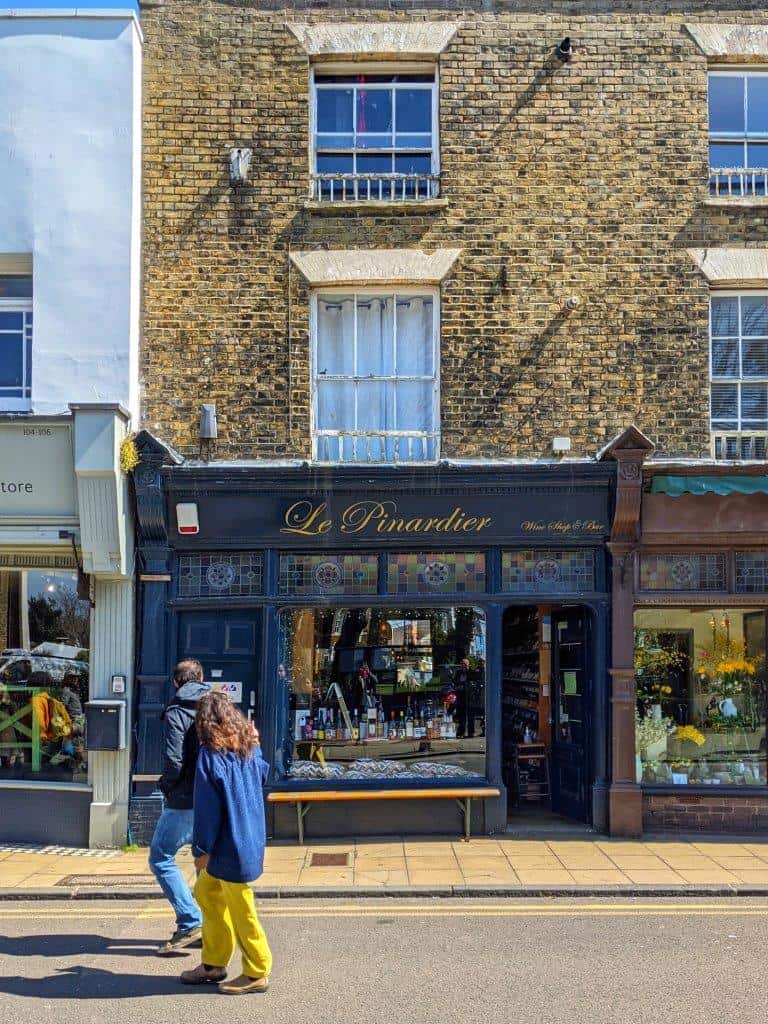 A handsome navy blue shop front called 'Le Pinardier' with bottles of wine in the window with two people looking in as they walk past