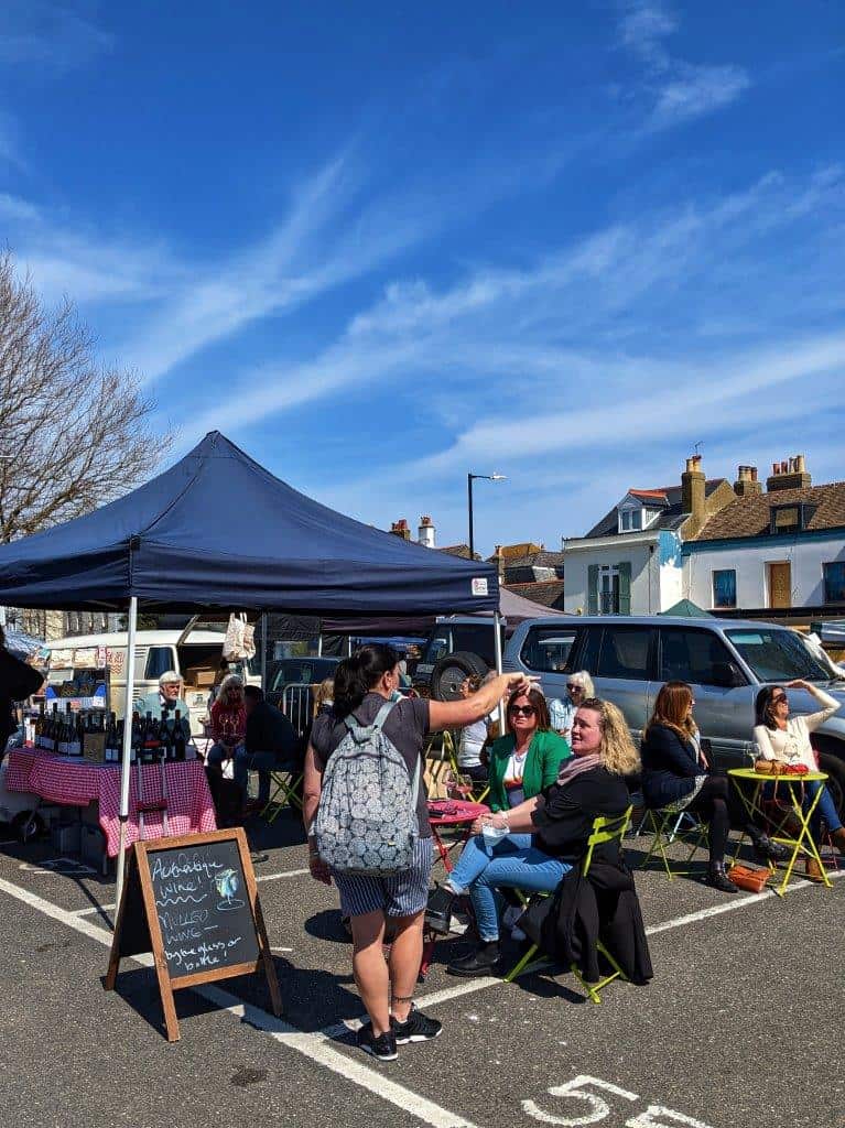 A gazeebo in a carpark under which there is a table laden with tine bottles, outside women sit on folding chairs enjoying their glasses of wine under a blue sky