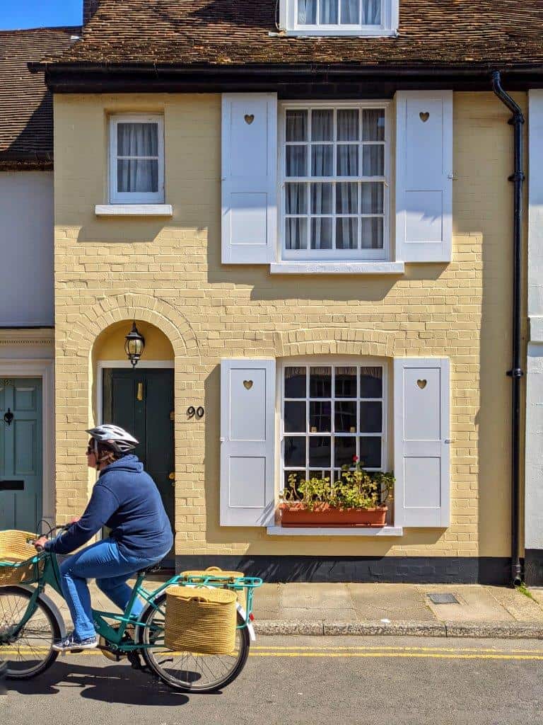 A buttercup yellow house front on with white window frames and shutters, a blue door and a person riding past it on a bicycle with baskets strapped to the back