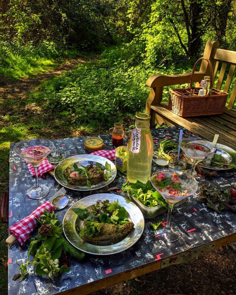 A bucolic picnic set-up laid out on a tressele table in a woodland clearing surrounded by foliage and a wooden bench, the table laden with springtime dishes, cutlery and drinks