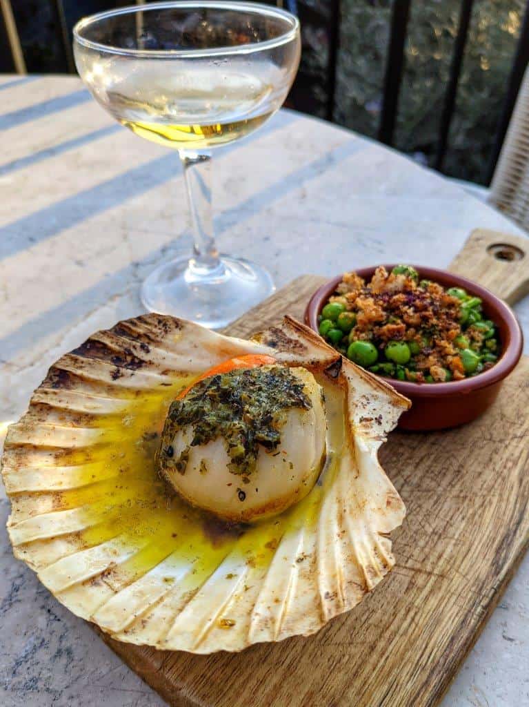 A restaurant dish of a cooked scallop served in its shell on a wooden board with a side of peas and a coupé glass of wine