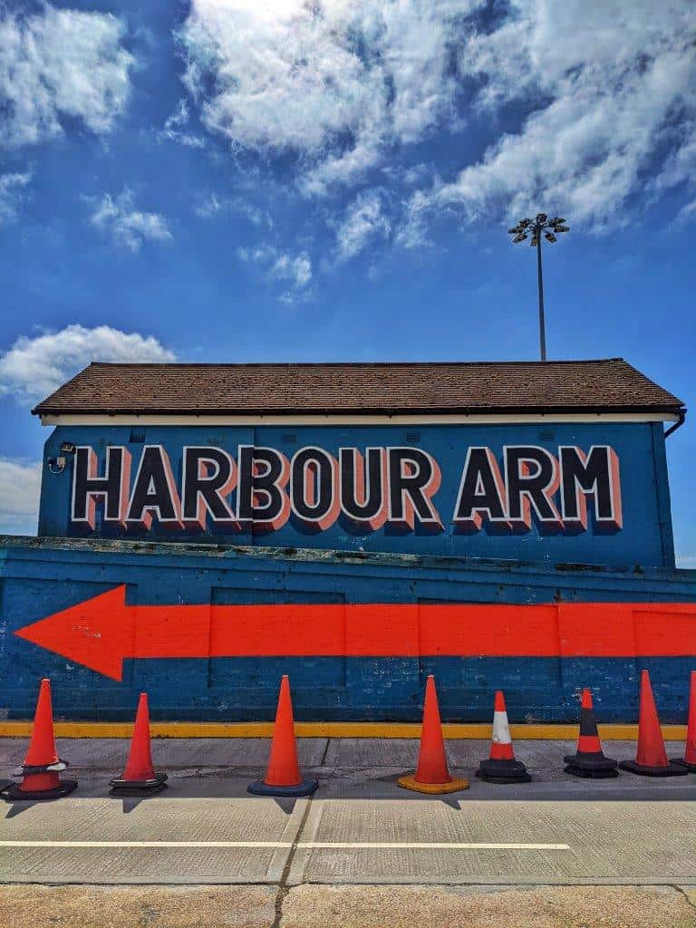 A small building painted blue with lettering that fills the whole of one wall reading 'Harbour Arm', underlined with a large red arrow pointing towards the left