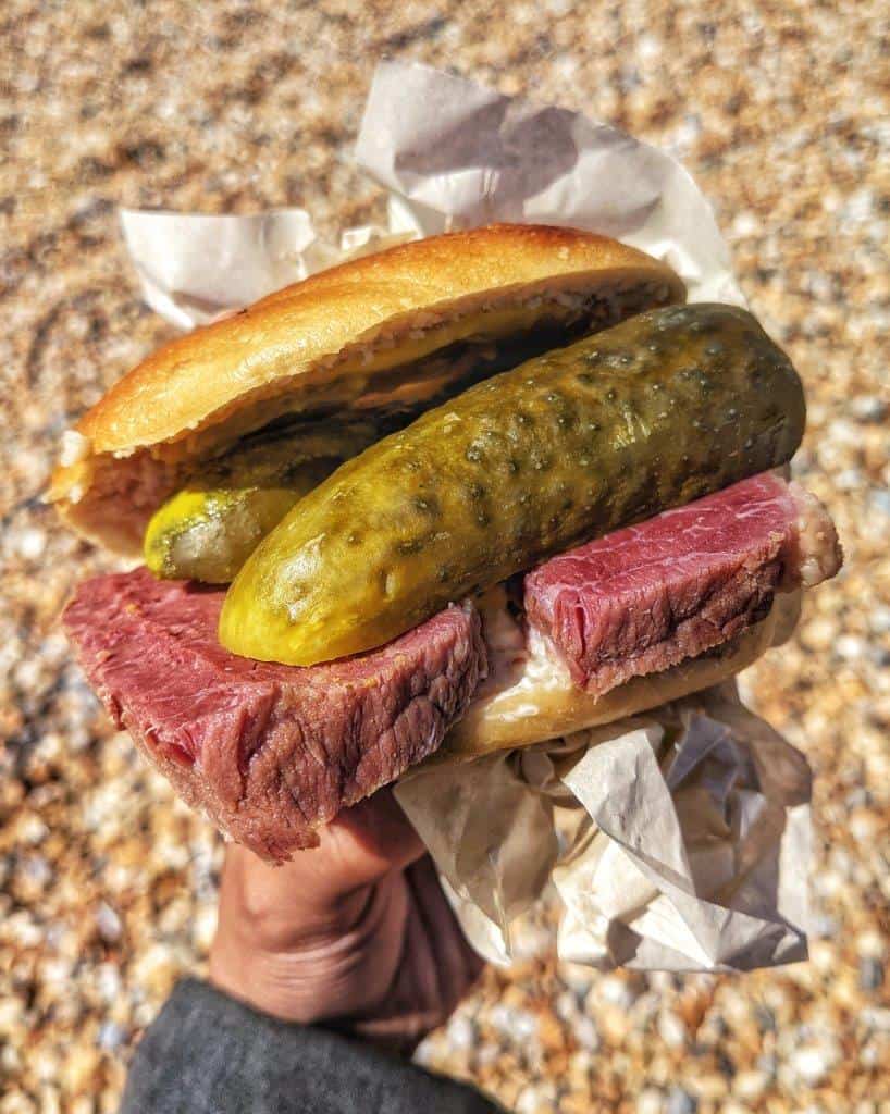 A close up of a hand holding a delicious bagel stuffed with thick slices of salt beef and huge pickled cucumbers, held over a pebbled beach