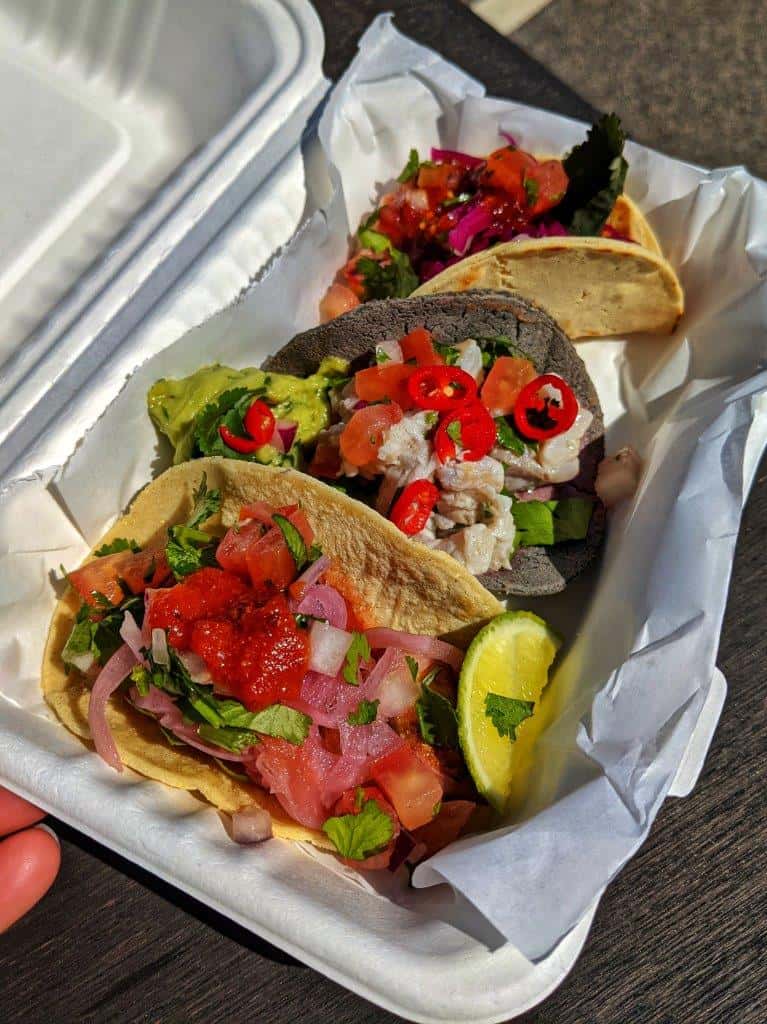 A close-up of a trio of colourful tacos in a takeaway box dressed with lime, tomatoes and coriander