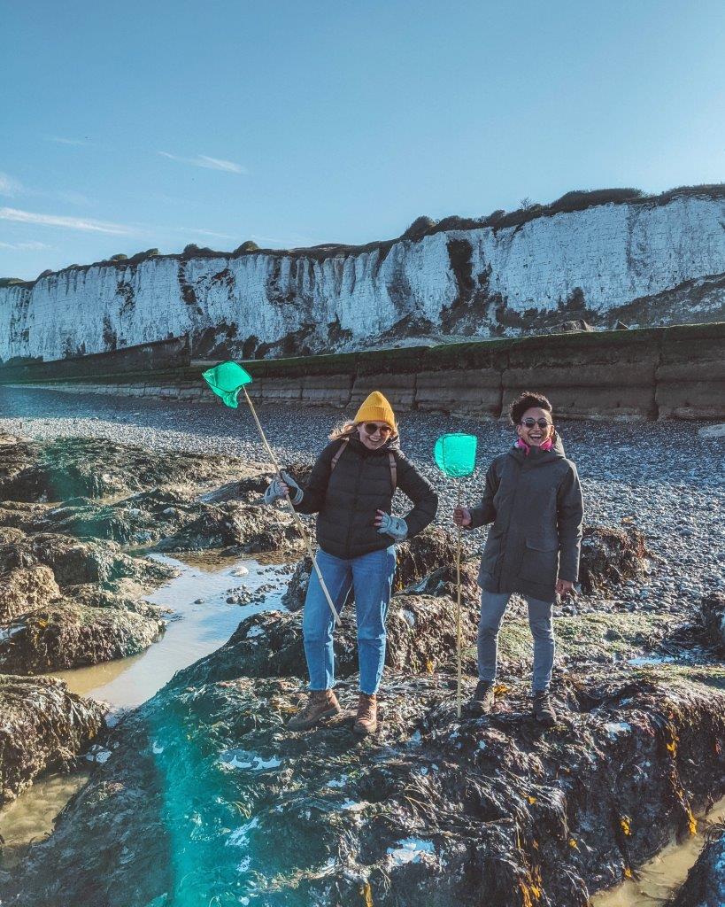 Two girls standing on big rocks on a pebble beach against a backdrop of white cliffs, both holding fishing nets and with big smiles