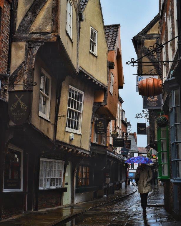 A woman holding a purple umbrella walking down a cobbled medieval looking street in the rain, with overhanging timber-framed buildings