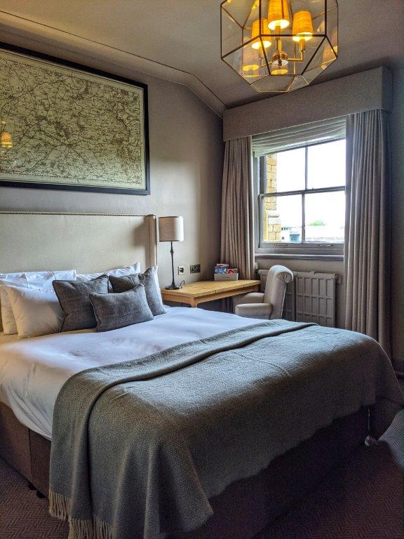 A hotel room with a large and freshly made double bed next to a small desk and chair with a window and heavy draped curtains