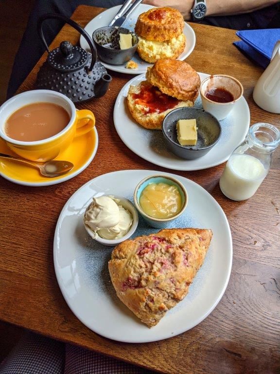 Plates of scones, preserves and clotted cream laid out on a wooden table along with cups and pots of tea