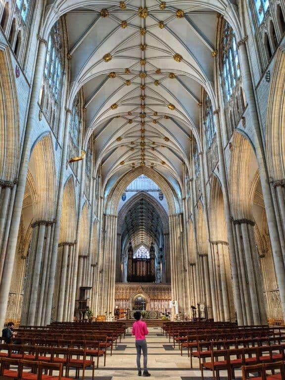 A view down the central aisle of a grand cathedral with a woman in a pink top standing in the middle, flanked by rows of pews, gazing up at the towering domed ceiling