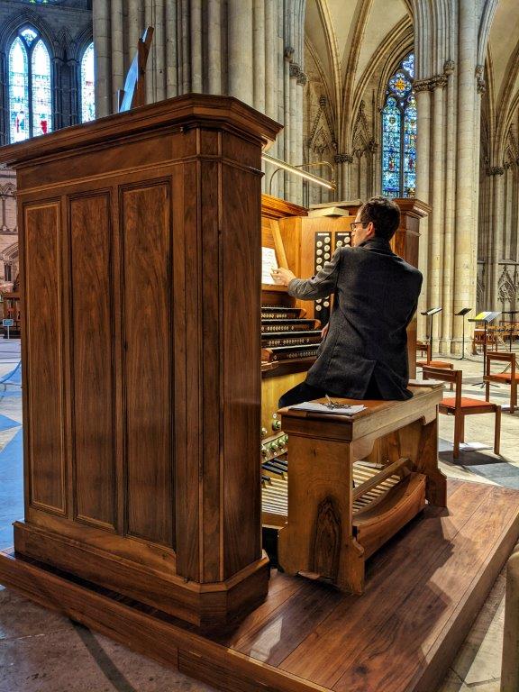 A man sitting on a large piano stool with his back to us, playing the organ in a grand church