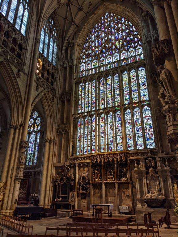 An imposing and intricate stained glass window reaching the full height of the cathedral, about the size of a tennis court. Some chairs in the foreground dwarfed by the great window's presence