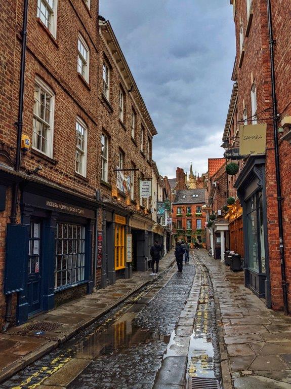 The view down a narrow cobbled street after the rain flanked by red brick industrial buildings housing independent shops on the ground floor