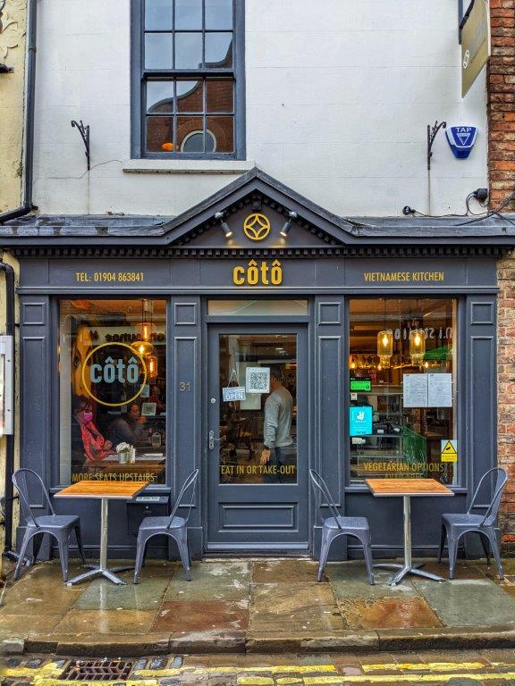 A dark grey shop front with yellow lettering reading 'Coto' and two tables out the front