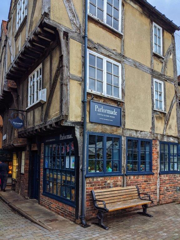 The corner view of a shop called 'Parlormade' in a medieval building with a wooden bench out the front and overhanging timber-framed upper stories