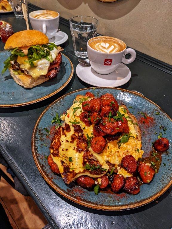 A blue plate loaded with scrambled eggs and chorizo alongside a stuffed breakfast bun and coffes, set out on a wooden breakfast bar