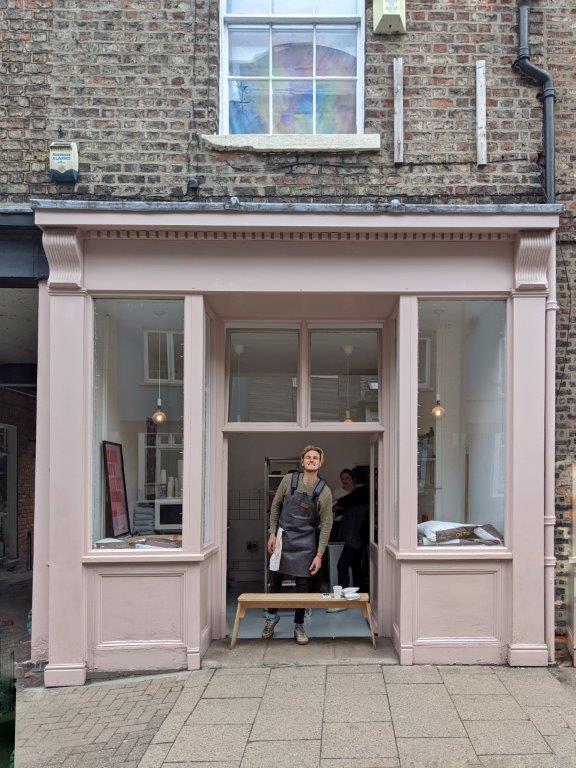 A blush pink shop front with no name, with a man wearing an apron standing in the entrance grinning to camera