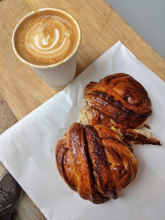 Two glossy buns laid on a paper bag and a coffee in a paper cup on a wooden bench