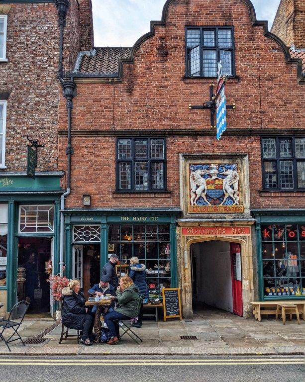 Three friends eating at a small table on the pavement in front of a shop called 'The Hairy Fig', part of a larger red brick building with an entrance to a courtyard to the right