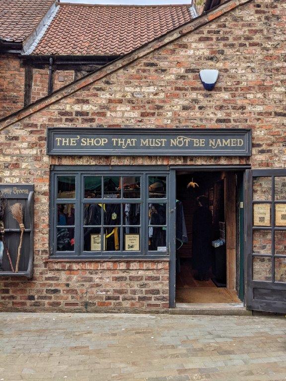 A dark grey and brick shop front called 'The Shop That Must Not Be Named' with the entrance door wide open