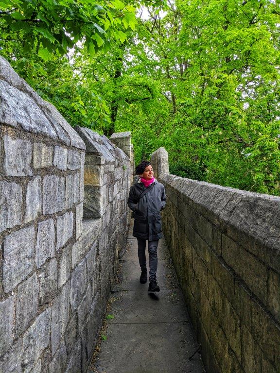 A woman in a coat and pink scarf striding towards the camera, peering over a stone wall to her left