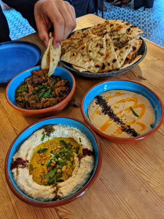 A spread of North African-inspired mezze dishes on colourful crockery, including a big pile of flat breads