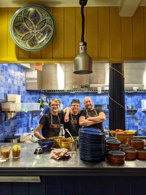 Three chefs standing behind the pass in a restaurant kitchen smiling to camera wearing visors