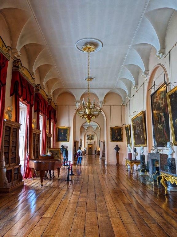 A grand hall with floor to ceiling windows, draped red velvet curtains, a chandelier and classic portraits hung on the walls