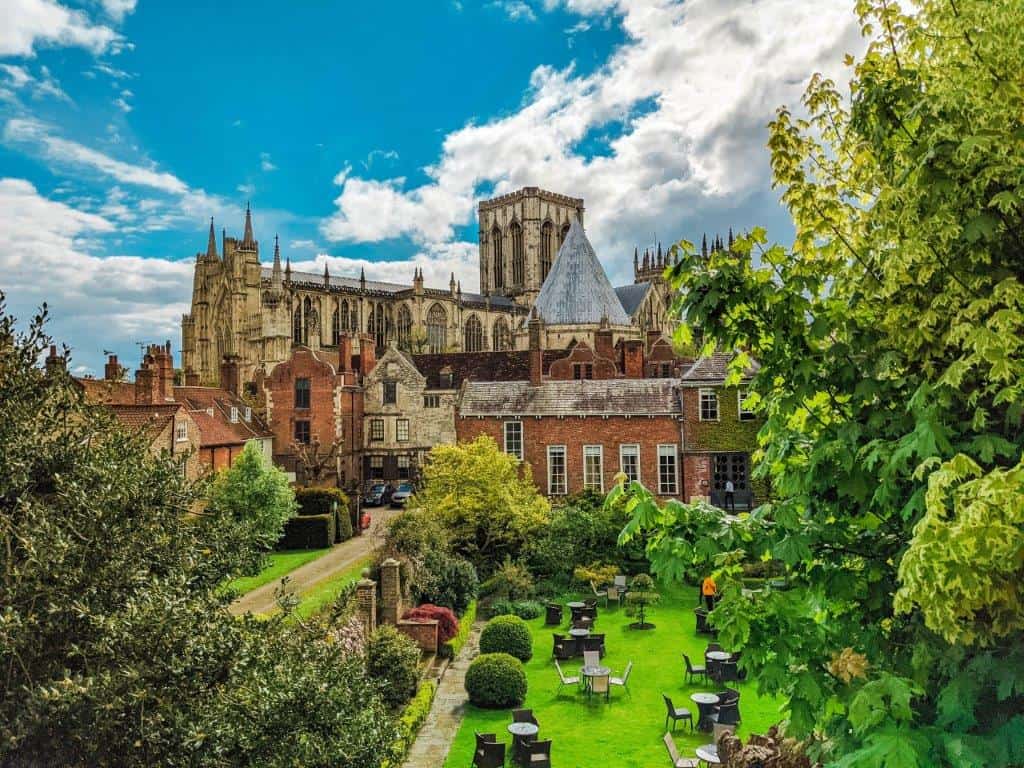 A landscape view of a grand cathedral on a sunny day, partially concealed by other brick buildings and tree foliage, with a well manicured lawn in the foreground