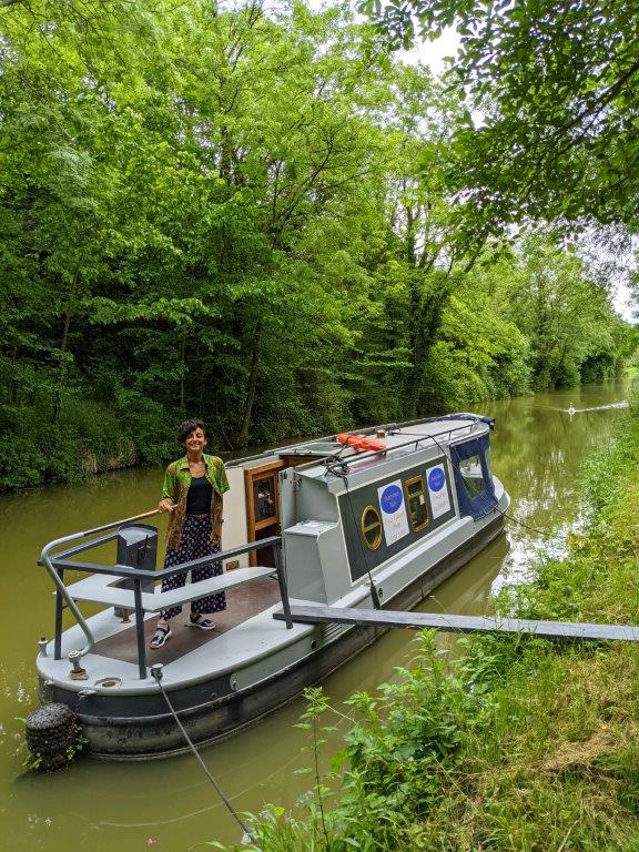 A narrowboat moored up on a canal with a wooden plank linking it to the bank and a woman standing on deck